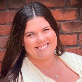 A smiling person with a white shirt with brick wall in the background